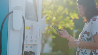 Genç, güzel Asyalı bir kadın elektrik istasyonunda duruyor, kablolu interneti kuruyor, akıllı telefondan internete bağlanıyor ve elektrikli araba ile seyahat ediyor.