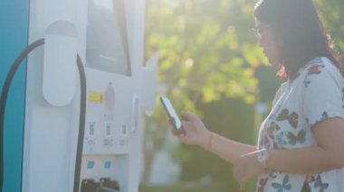 Genç, güzel Asyalı bir kadın elektrik istasyonunda duruyor, kablolu interneti kuruyor, akıllı telefondan internete bağlanıyor ve elektrikli araba ile seyahat ediyor.