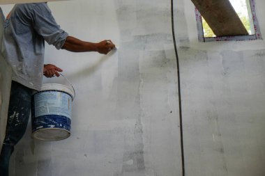 Asyalı mekanik ressam, inşaat alanının içindeki beton duvara beyaz boya yapmak için fırça kullanıyor.