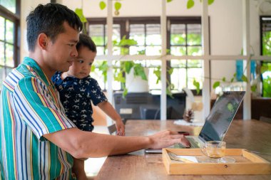 Asyalı baba internette bilgisayarla çalışıyor. Bebek bebekle kahve dükkanında oturuyor. 