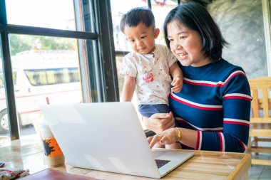 İnternette iş yapan Asyalı bir anne kucağında dizüstü bilgisayarla çalışıyor.