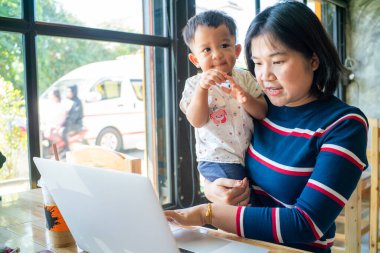 İnternette iş yapan Asyalı bir anne kucağında dizüstü bilgisayarla çalışıyor.