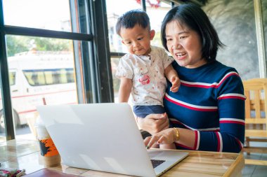 İnternette iş yapan Asyalı bir anne kucağında dizüstü bilgisayarla çalışıyor.