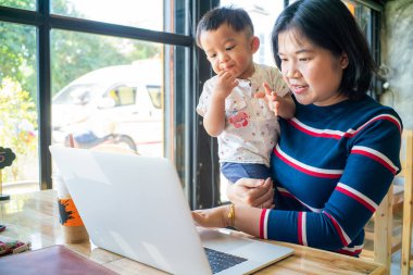 İnternette iş yapan Asyalı bir anne kucağında dizüstü bilgisayarla çalışıyor.