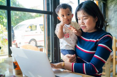İnternette iş yapan Asyalı bir anne kucağında dizüstü bilgisayarla çalışıyor.