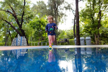 Küçük anaokulu 3 yaşındaki kız yağmurlu bir günden sonra oyun parkında suyun üzerinde koşuyor. 