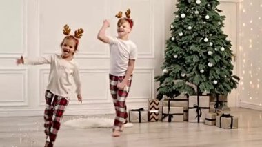 Two cheerful children are running around the living room in their pajamas near the Christmas tree.
