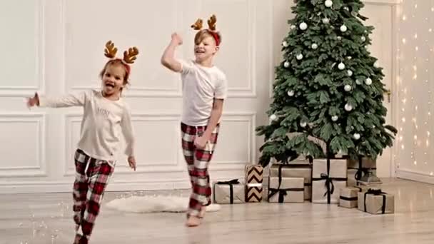 两个快乐的孩子穿着睡衣在圣诞树旁的客厅里跑来跑去 — 图库视频影像