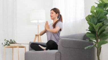 Yoga exercise concept, Young Asian woman doing yoga exercise in lotus pose with namaste on couch.