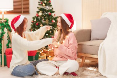 Noel kutlaması konsepti, Noel Baba şapkalı genç Asyalı kadın arkadaşının üzerine yılbaşı ışıkları süsler..