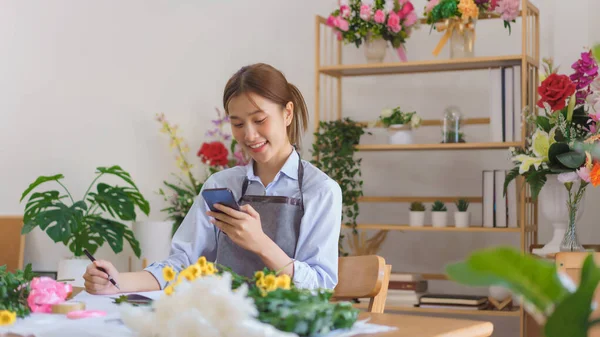 stock image Floristry concept, Woman florist look data on smartphone to taking notes in notebook at flower shop.