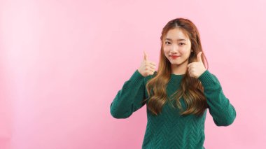 Genç kadınlar başparmaklarını her iki elinle kaldırır ve pembe arka planda boş bir alanla gülümserler..