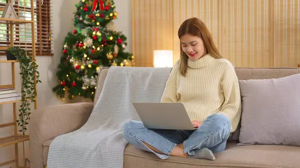 クリスマスの休日の概念 女性はクリスマスの木の近くのソファーに座っている間ノートパソコンで働いています ストック画像