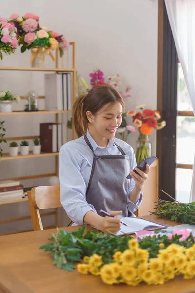 Florist concept, Woman florist looks data on smartphone to taking notes in notebook at flower shop.