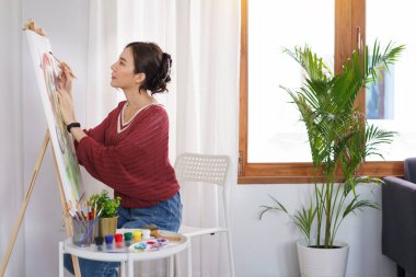Sanat konsepti, genç Asyalı kadın sandalyede oturur ve tuvale boya fırçası kullanır..