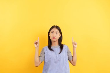 Young women points with finger up and doubtful face with empty space on isolated yellow background.