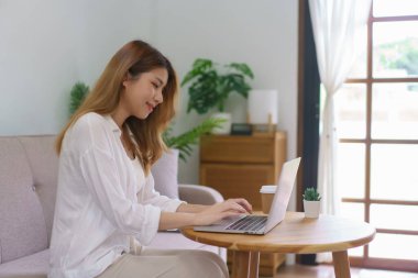 Ev konseptinden çalış, genç Asyalı kadın bilgisayarı kullanarak veri yaz ve oturma odasında çalış..