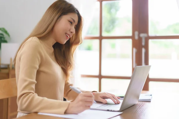 stock image Work from home concept, Young Asian woman reading data on laptop and taking notes on notebook.