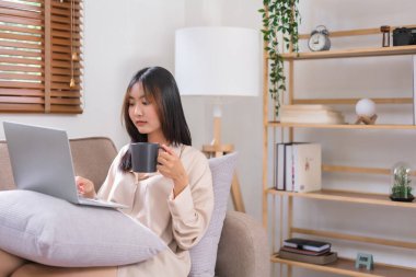Evde rahatlama kavramı, dizüstü bilgisayarla sosyal medyada gezinen ve kahve içen genç Asyalı kadın..