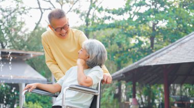 Yaşlı adam engelli bir kadının tekerlekli sandalyesini parkta yürümeye ve rahatlamaya itiyor..