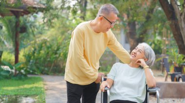 Yaşlı adam engelli bir kadının tekerlekli sandalyesini parkta yürümeye ve rahatlamaya itiyor..