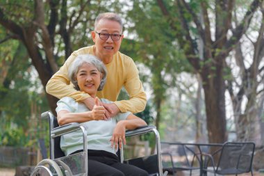 Kıdemli adam tekerlekli sandalyedeki engelli karısını kucaklıyor ve parkta mutlulukla baş parmağını gösteriyor..