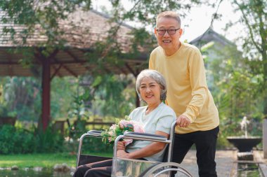 Yaşlı adam, sakat bir kadının tekerlekli sandalyesini çiçek buketiyle parkta yürümeye ve dinlenmeye itiyor..