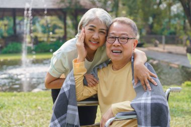 Parkta dinlenirken tekerlekli sandalyedeki engelli kocayı battaniyeyle kucaklayan yaşlı kadın..