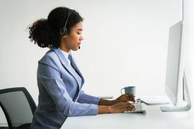 Kulaklıklı Afrikalı Amerikalı kadın bilgisayar üzerinde çalışıyor ve çağrı merkezinde müşteri desteği hakkında konuşuyor..
