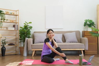 Sportif kadın yoga minderinde oturur ve evde egzersiz yapmak için internetten yoga eğitimini izledikten sonra gülümser..