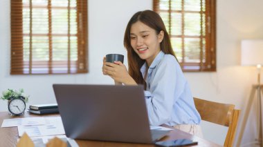 Business at home concept, Businesswoman reading marketing plan data on laptop and drinking coffee.