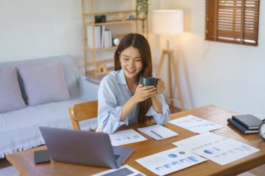 Business at home concept, Businesswoman reading marketing plan data on laptop and drinking coffee.