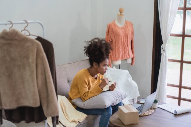 Afrikalı Amerikalı moda tüccarı kadın dizüstü bilgisayardan online siparişleri kontrol ediyor ve deftere veri yazıyor..