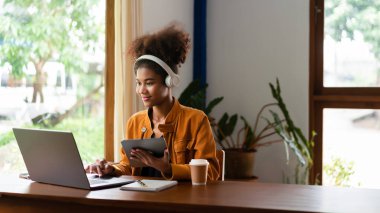 Afro-Amerikalı iş kadını kafede tablet üzerinde çalışıyor ve iş verilerini laptopta yazıyor..