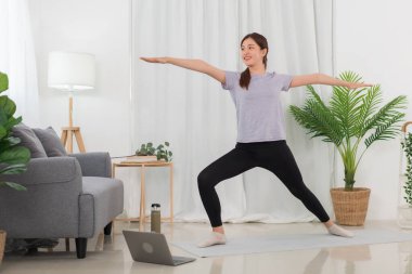 Yoga exercise concept, Young Asian woman watching yoga tutorial online to doing warrior pose on mat.