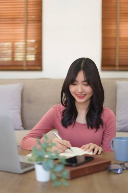 Genç iş kadını laptopta veri okuyor ve evde çalışırken deftere notlar yazıyor.. 