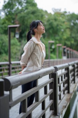 Genç Asyalı koşucu kadınlar gölün yanında durup kulaklık takarak parkta antrenman yaptıktan sonra dinlenmek ve sağlıklı yaşam tarzı için spor yapmak için kulaklık takıyorlar..