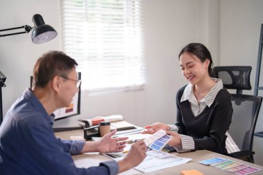 Interior designer women explaining blueprint of construction architectural and color swatch samples for interior design to senior businessman while discussing to designing about building of project.
