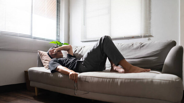 Человек с психическими проблемами лежит на диване и с помощью руки, чтобы покрыть глаза депрессивной эмоцией.