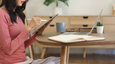 Genç iş kadını tablete iş projesi hakkında notlar yazıyor ve evde dizüstü bilgisayar üzerinde çalışıyor..