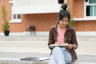 Modern bir okul binasının önünde oturan bir lise öğrencisi deftere bir şeyler yazıyor. Kahverengi ceket ve açık mavi kot giyiyor. Yazılarına odaklanmış.. 