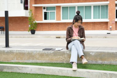 Modern bir okul binasının önünde oturan bir lise öğrencisi deftere bir şeyler yazıyor. Kahverengi ceket ve açık mavi kot giyiyor. Yazılarına odaklanmış.. 