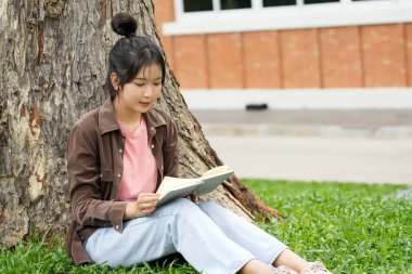 Modern bir okul binasının önünde oturan bir lise öğrencisi deftere bir şeyler yazıyor. Kahverengi ceket ve açık mavi kot giyiyor. Yazılarına odaklanmış.. 