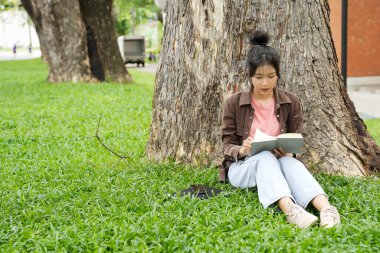 Modern bir okul binasının önünde oturan bir lise öğrencisi deftere bir şeyler yazıyor. Kahverengi ceket ve açık mavi kot giyiyor. Yazılarına odaklanmış.. 