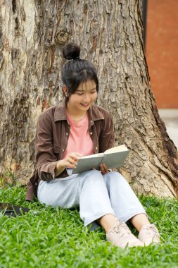 Modern bir okul binasının önünde oturan bir lise öğrencisi deftere bir şeyler yazıyor. Kahverengi ceket ve açık mavi kot giyiyor. Yazılarına odaklanmış.. 