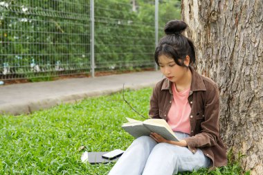 Modern bir okul binasının önünde oturan bir lise öğrencisi deftere bir şeyler yazıyor. Kahverengi ceket ve açık mavi kot giyiyor. Yazılarına odaklanmış.. 