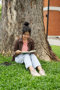Modern bir okul binasının önünde oturan bir lise öğrencisi deftere bir şeyler yazıyor. Kahverengi ceket ve açık mavi kot giyiyor. Yazılarına odaklanmış.. 