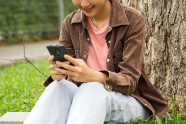 Bir lise öğrencisi bir ağacın altındaki çimlerin üzerinde oturuyor, akıllı telefonuna bakarken gülümsüyor. Kahverengi bir ceket ve açık mavi kot pantolon giyiyor, rahat ve mutlu görünüyor.. 