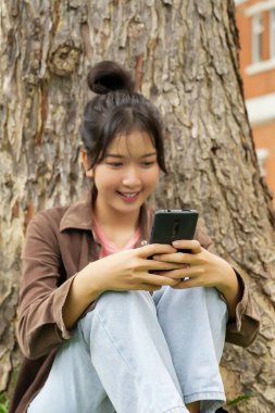 Bir lise öğrencisi bir ağacın altındaki çimlerin üzerinde oturuyor, akıllı telefonuna bakarken gülümsüyor. Kahverengi bir ceket ve açık mavi kot pantolon giyiyor, rahat ve mutlu görünüyor.. 