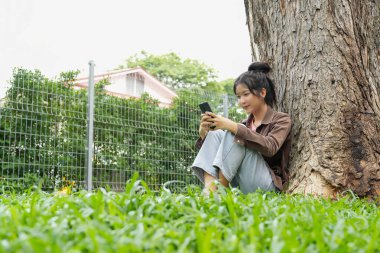 Bir lise öğrencisi bir ağacın altındaki çimlerin üzerinde oturuyor, akıllı telefonuna bakarken gülümsüyor. Kahverengi bir ceket ve açık mavi kot pantolon giyiyor, rahat ve mutlu görünüyor.. 